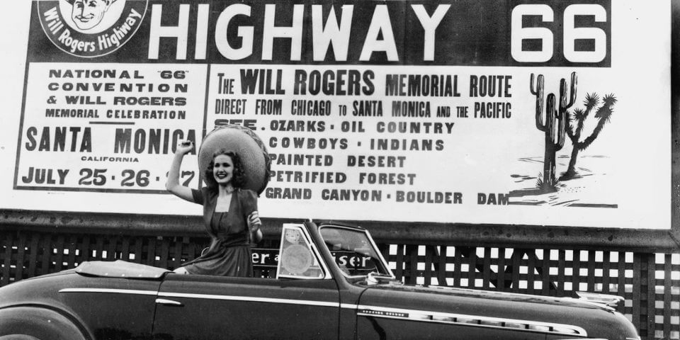 A woman sits on the backseat of an old car in front of the Highway 66 sign.