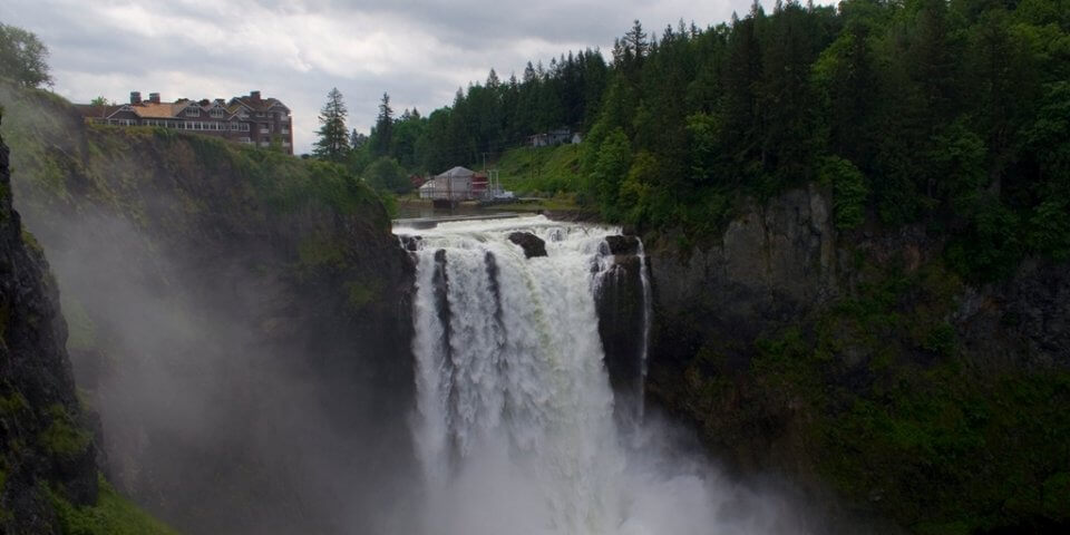 Snoqualmie Falls in Washington on a cloudy day. A hotel is at the top of it as well as a lot of thick coniferous trees.