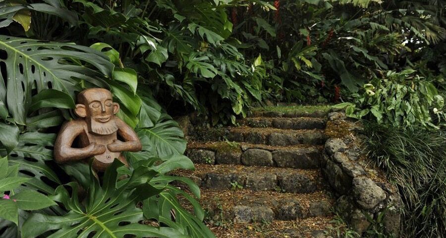 A wooden sculpture of a Menehune resting his hands on his belly next to a stone path in the Hawaiian forest.