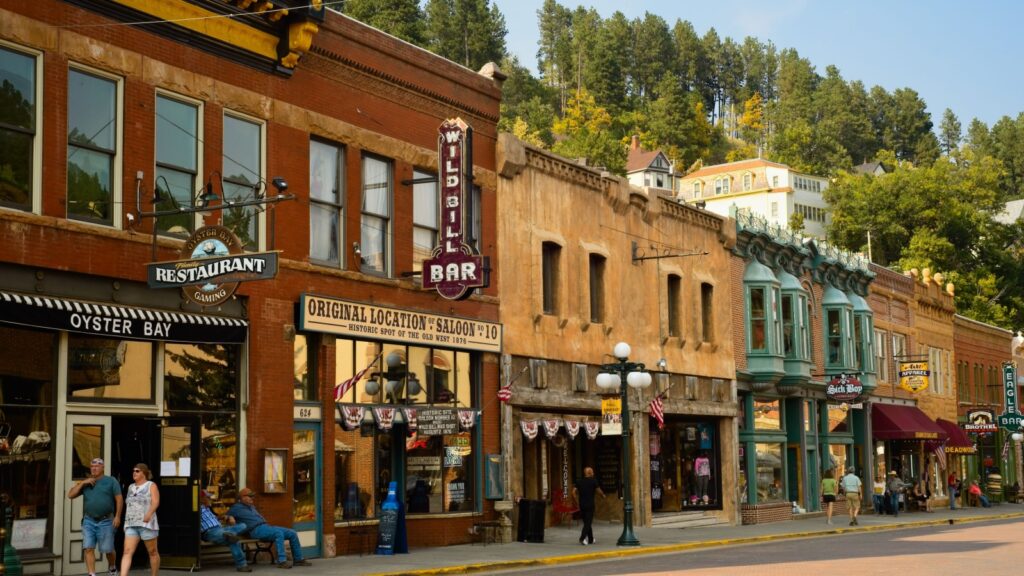 A street scene in Deadwood, South Dakota, featuring historic buildings with businesses like "Oyster Bay Restaurant," "Wild Bill Bar," and "Original Location Saloon #10" lining the street, with people walking on the sidewalk.