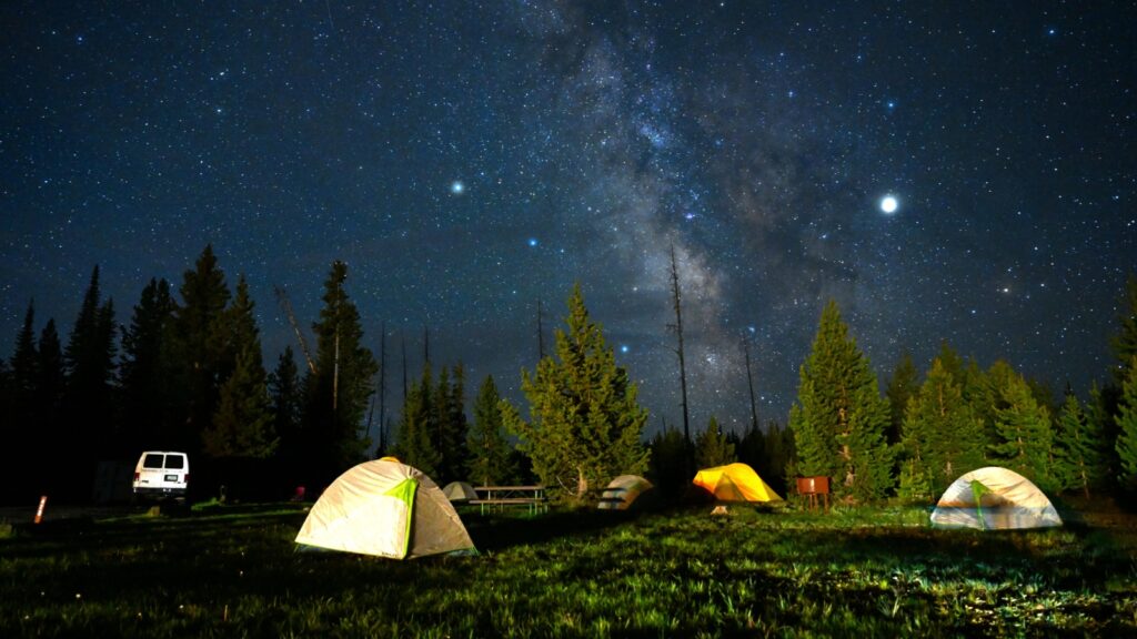 Illuminated tents under a starry night sky with the Milky Way visible over a forest in Yellowstone National Park, Wyoming.