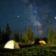 Illuminated tents under a starry night sky with the Milky Way visible over a forest in Yellowstone National Park, Wyoming.