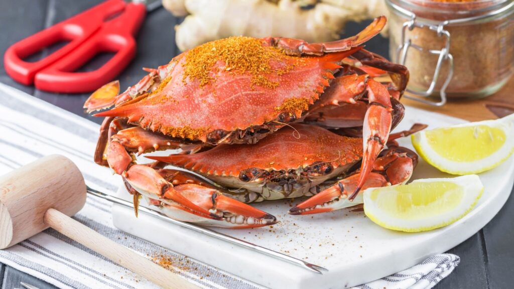Two cooked blue crabs seasoned with Old Bay, served on a white platter with lemon wedges, a wooden mallet, and red crab crackers, alongside ginger and a spice jar in the background.