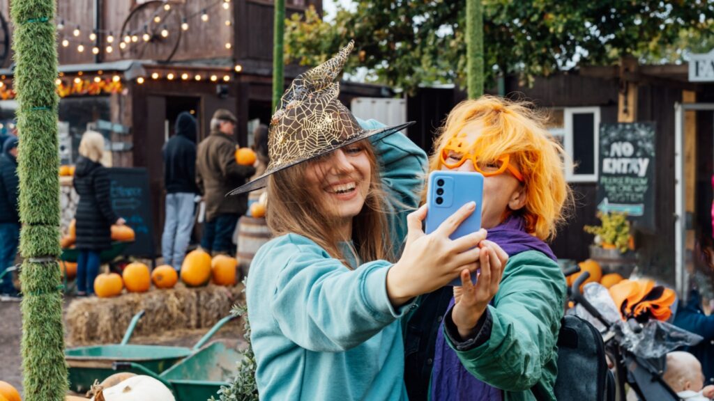 Two laughing female friends in fun glasses and witch hat having fun and making selfie on phone on decorated pumpkin farm.
