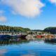 A scenic view of Lake Cumberland featuring a marina with numerous boats docked along the shore, backed by lush green hills under a bright blue sky with scattered clouds.