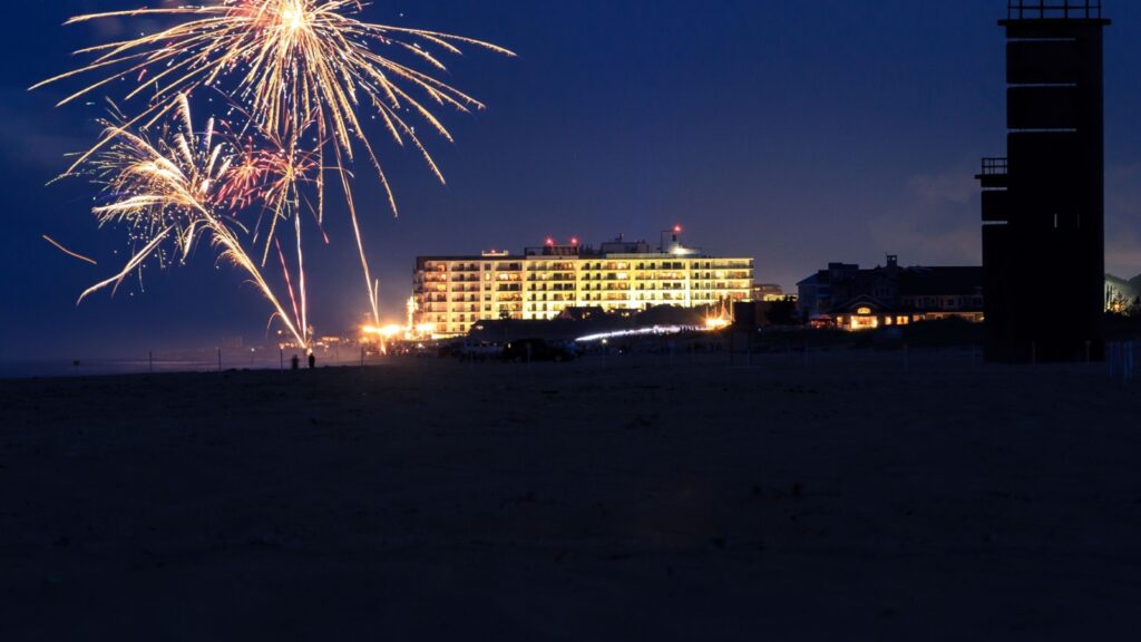 Fireworks illuminate the night sky over Rehoboth Beach during a jazz festival, with a brightly lit building and a tall structure visible in the background.
