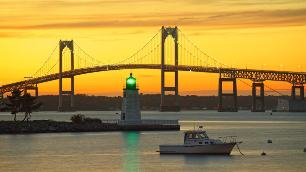 A sunset view of the Newport Bridge and Goat Island Lighthouse in Newport, Rhode Island, with a boat anchored in the foreground.
