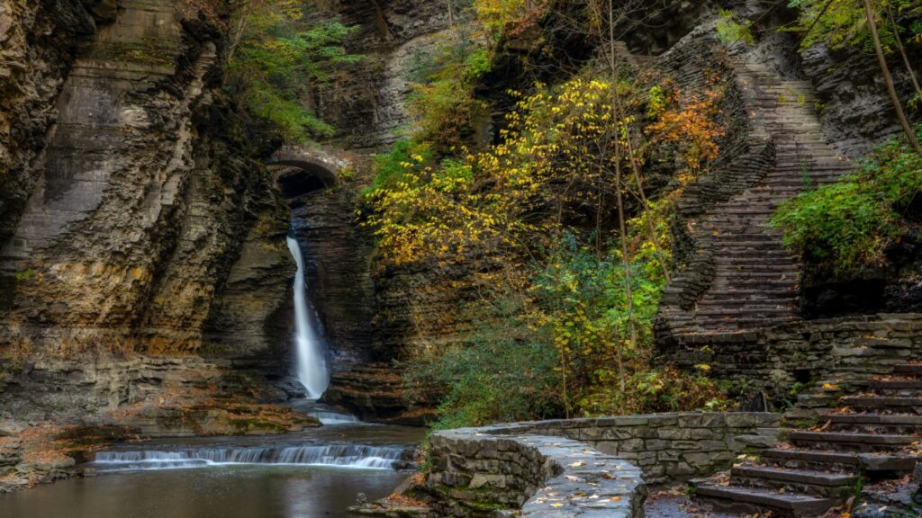 A scenic view of the Gorge Trail at Watkins Glen State Park, featuring a waterfall cascading into a pool, a stone path winding along the gorge, and lush autumn foliage on the surrounding cliffs.