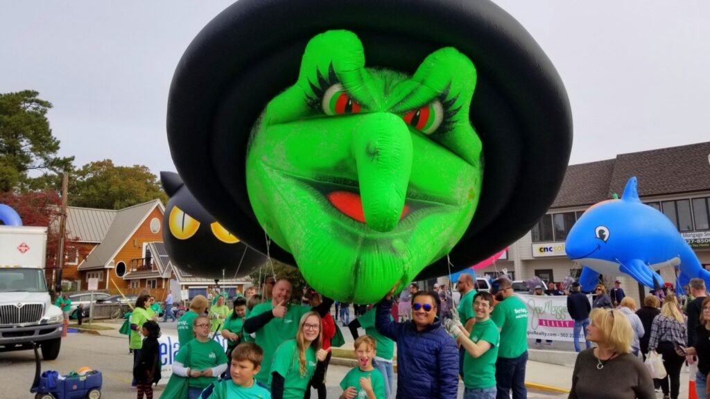 A large inflatable green witch float, a blue dolphin float, and people in green shirts participating in the Sea Witch Festival parade in Rehoboth Beach.