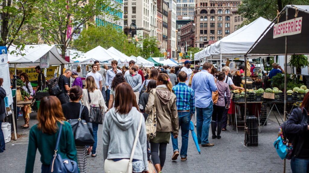 A bustling outdoor farmers market with numerous people browsing produce stalls and tents in an urban setting, likely the Union Square Greenmarket in New York City.