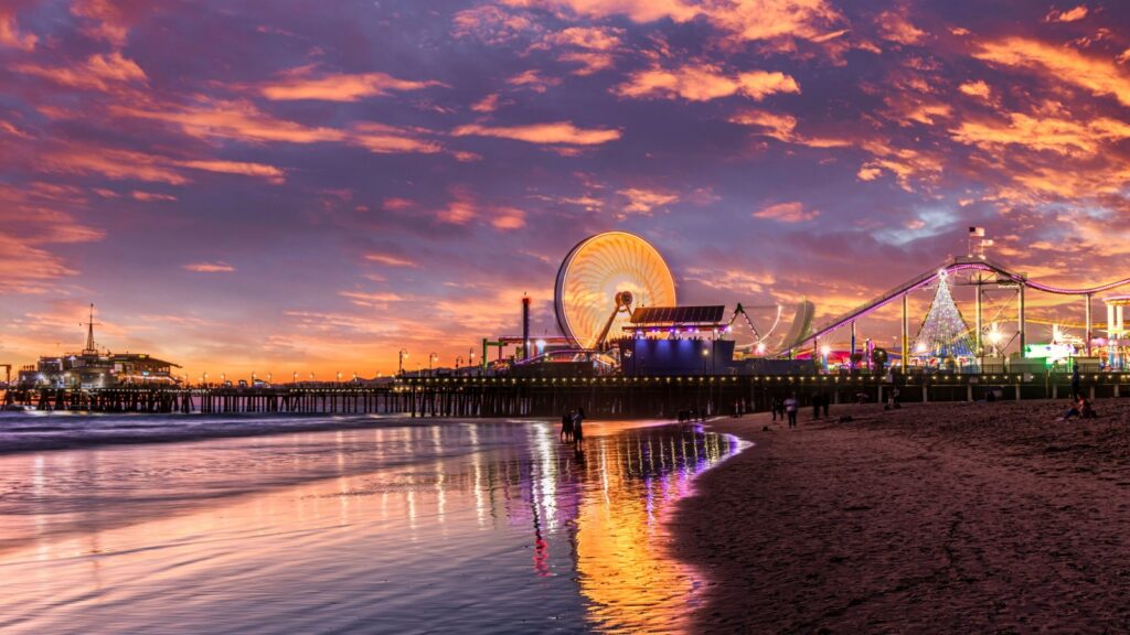A vibrant sunset view of Santa Monica Pier in California, featuring its illuminated Ferris wheel, roller coaster, and colorful reflections on the wet sand along the shoreline.