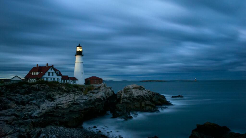 Portland Head Light in Cape Elizabeth, Maine, illuminated at dusk or night with a cloudy sky and rocky coastline. 