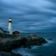 Portland Head Light in Cape Elizabeth, Maine, illuminated at dusk or night with a cloudy sky and rocky coastline.