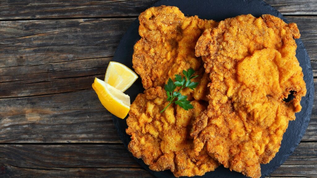 Golden-brown Wiener schnitzel with lemon and parsley on a dark plate.
